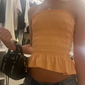 Orange smockad bandeau tubtopp - Superfin orange bandeau tubtopp med smockad överdel och volangkant nertill. Toppen är ärmlös och har en söt, somrig vibe som passar perfekt till varmare dagar.
