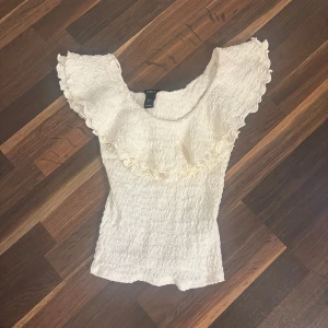 Vit volangtopp från Lindex XS - Supersöt vit topp från Lindex i storlek XS. Toppen har volangdetaljer över axlarna och bröstet, samt ett vågigt, ribbat tyg som ger en unik struktur.