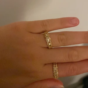 Guldiga ringar med glittriga stenar - Två snygga guldiga ringar med rektangulära glittriga stenar runt hela ringen. Perfekt för dig som vill ha lite extra bling på fingrarna. Ringen har en lyxig känsla och passar till alla tillfällen när du vill sticka ut.