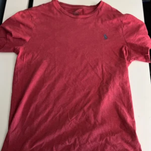 Röd t-shirt från Polo Ralph Lauren - Röd t-shirt från Polo Ralph Lauren med klassisk rund hals och korta ärmar. T-shirten har en broderad blå logga på bröstet och är gjord i mjuk bomull. Perfekt basic-plagg med snygg passform och stilren look.