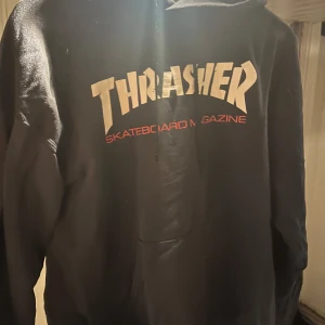 Svart Thrasher hoodie med tryck - Svart hoodie från Thrasher med klassiskt stort logotryck i vitt och rött på bröstet. Tröjan har huva och känguruficka framtill, perfekt för en streetwear-look. Mjuk insida och avslappnad passform. Osäker på storlek men mig som oftast är en storlek M/L