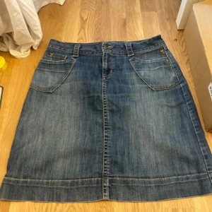 Jeanskjol från Time, modell Karin - Klassisk blå jeanskjol från Time, modell Karin. Kjolen har fem fickor, bälteshällor och stängs med knapp och dragkedja. Snygg, rak passform och i tåligt jeanstyg. Perfekt till sneakers eller boots för en chill look.