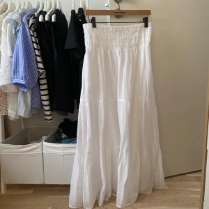 Vit långkjol med smock i midjan - Superfin vit långkjol med smockad midja och lätt, flowigt tyg. Kjolen ger en luftig känsla, perfekt för varma dagar. Enkel att matcha med både toppar och t-shirts för en chill look.Endast använd 1 gång 