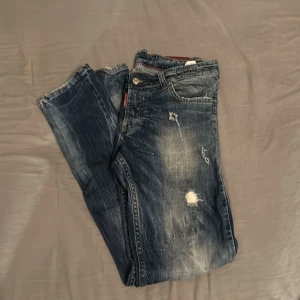 Dsquared2 blå distressed jeans - Snygga blå jeans från Dsquared2 med coolt distressed och slitna detaljer samt färgstänk. Klassisk femficksmodell med läderpatch bak i midjan och Dsquared2-logga vid fickan. Jeansen har en rak passform och är tillverkade i bomull.