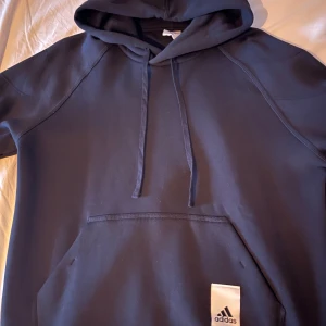 Svart hoodie från Adidas med huva - Svart hoodie från Adidas i storlek M. Klassisk modell med huva, snörning och stor magficka framtill. Adidas-logga på fickan. Perfekt för chill eller träning, tillverkad i mjukt material som känns skönt mot huden. Aldrig använts men ingen prislapp kvar tyvärr. Nypris 799kr!