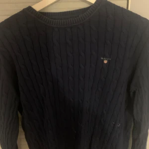 Mörkblå kabelstickad tröja från Gant - Kabelstickad mörkblå tröja från Gant med rund halsringning och diskret logga broderad på bröstet. Klassisk och stilren modell med långa ärmar, perfekt för dig som gillar preppy vibes. Tillverkad i mjukt material som känns skönt mot huden.