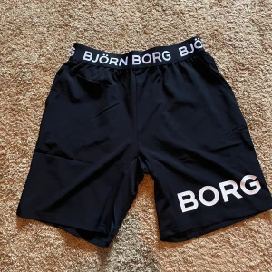 Svarta Björn Borg shorts i M - Svarta shorts från Björn Borg med bred resår i midjan där loggan står tryckt runtom. Stora vita BORG-bokstäver på ena benet. Perfekta för träning, tillverkade i ett lätt och smidigt syntetmaterial som andas. PRISLAPP KVAR! Aldrig använda! Nypris 399kr! 