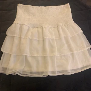 Vit volangkjol från BikBok - Supersöt vit kortkjol från BikBok med flera lager volanger och smockad midja. Kjolen är luftig och har en romantisk vibe, perfekt för varma dagar. Materialet känns lätt och flowy, vilket gör den extra bekväm att bära.