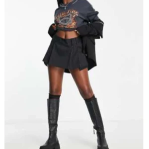 Svart randig kortkjol med veck - Snygg svart kortkjol med tunna vertikala ränder och veckad design. Kjolen har en modern och edgy look som passar perfekt till boots och oversized jacka. Enkel att styla för en cool streetstyle.