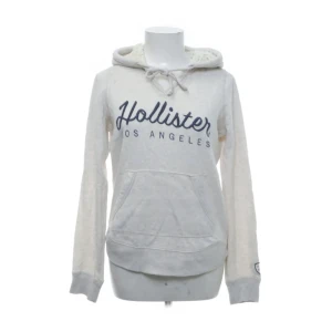 Vit hoodie från Hollister med tryck - Mysig vit hoodie från Hollister med mörkblått tryck 'Hollister Los Angeles' på bröstet. Tröjan har huva med snörning, känguruficka och mjukt foder i luvan. Perfekt för chill dagar och enkel att matcha med jeans eller leggings.