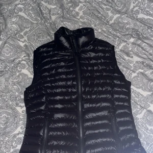 Svart dunväst med dragkedja - Snygg svart dunväst med hög krage och horisontella quiltade sömmar. Västen har två fickor med dragkedja framtill och en stilren, glansig finish. Perfekt att slänga över en hoodie eller tröja för extra värme.