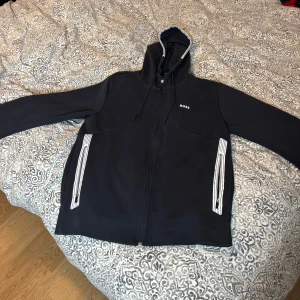 Zip up från BOSS med huva - Mörkblå zip up från BOSS med dragkedja, huva med snörning och vita detaljer vid fickorna. Tröjan har en clean look med diskret BOSS-logga på bröstet och passar perfekt för dig som gillar stilren streetwear.