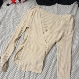 Beige ribbad omlottopp med v-ringning - Snygg beige ribbad topp med omlott och djup v-ringning. Långärmad och figurnära passform, perfekt att styla med jeans eller kjol. Mjuk och stretchig, passar både till lager-på-lager och som den är.