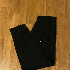 Svarta Nike Therma-Fit mjukisbyxor - Svarta mjukisbyxor från Nike med Therma-Fit-teknologi. Byxorna har elastisk midja och en vit Nike-logga på benet. Perfekta för chill eller träning, riktigt sköna och stilrena.