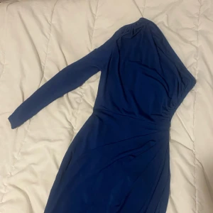 Marinblå långklänning one shoulder - Snygg marinblå långklänning med en ärm och one shoulder-design. Klänningen har draperade detaljer och en elegant, figurnära passform. Perfekt för dig som vill sticka ut med en stilren och modern look.