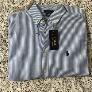 Blåvit randig skjorta Polo Ralph Lauren - Klassisk blå och vit randig skjorta från Polo Ralph Lauren med button-down krage och broderad logga på bröstet. Skjortan har lång ärm, knappar framtill och är tillverkad i mjuk bomull. Perfekt för en clean och stilren look. Ej använd 