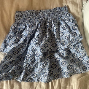 Blåvit mönstrad kortkjol med resår - Supersöt kortkjol i blått och vitt med ett unikt grafiskt mönster. Kjolen har bred smockresår i midjan som ger en skön och flexibel passform. Tillverkad i tunn och luftig bomull, perfekt för varma dagar. Passar dig som gillar en avslappnad och trendig stil.