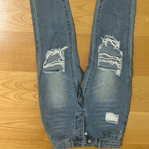 Blå ripped jeans med raka ben - Säljer ett par ljusblå jeans med raka ben och stora slitningar på båda knäna. Jeansen har klassisk femficksdesign, knappgylf och coolt sliten look. Perfekta för dig som gillar streetstyle och vill ha en avslappnad vibe.