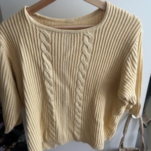 Beige stickad tröja med flätmönster - Säljer en beige stickad tröja med snygga flätade detaljer framtill och ribbad struktur. Tröjan har rund halsringning och trekvartsärmar, perfekt för lager-på-lager-stil. Passar dig som gillar en chill och klassisk look.