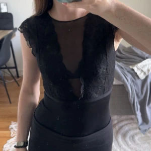 Svart spets body med öppen rygg - Säljer en svart body med transparent mesh och spetsdetaljer framtill och på axlarna. Djup v-ringning och öppen rygg ger en riktigt snygg look. Ärmlös med små spetsärmar och figurnära passform. Perfekt för dig som gillar trendiga och feminina plagg.