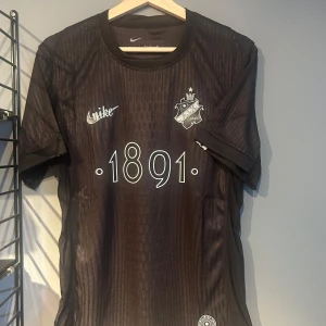 Svart AIK fotbollströja Nike 1891 - Snygg svart AIK fotbollströja från Nike med klubbmärke, trycket '1891' på bröstet och diskret mönster i tyget. Kortärmad modell i lätt och ventilerande material, perfekt för match eller läktaren. Texten 'SMOKINGLIRARE' i nacken. Helt ny med tagg en replika 