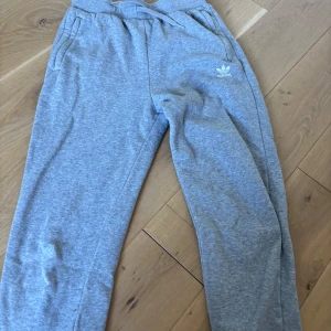 Grå mjukisbyxor från Adidas - Gråa mjukisbyxor från Adidas med klassisk logga på ena benet. Byxorna har dragsko i midjan, sidofickor och är tillverkade i mjukt bomullsmaterial. Perfekta för chill eller träning, med en relaxed vibe och enkel stil.