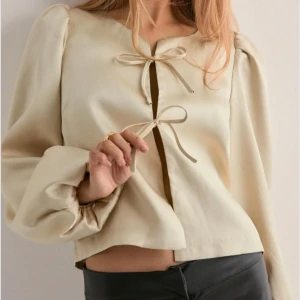 Beige blus med knytband - Stilren beige blus med långa puffärmar och två söta knytband framtill. Blusen har en croppad passform och är tillverkad i ett mjukt, lätt material som ger en lyxig känsla. Perfekt för dig som gillar en enkel men trendig look.