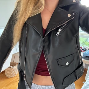 Svart skinnjacka från Zara med bälte - Säljer en svart skinnjacka från Zara i bikerstil med silvriga dragkedjor, fickor och bälte i midjan. Jackan har klassisk krage, långa ärmar och coola detaljer som ger en edgy vibe. Perfekt för dig som vill ha en tidlös och snygg look.