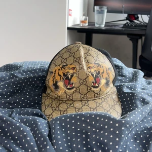 Gucci keps med tigertryck och GG-mönster - Säljer en ikonisk Gucci keps i beige med klassiskt GG-monogram och två stora tigerhuvuden framtill. Kepsen har svart mesh baktill och justerbart band med gröna och röda ränder. Perfekt statement-piece för dig som vill sticka ut.