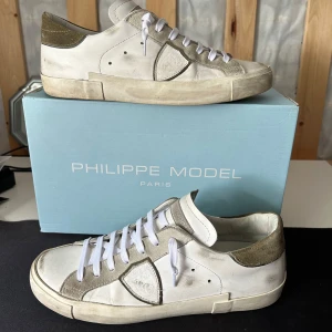 Philippe Model  - Snygga vita sneakers från Philippe Model med beige mockadetaljer och vita snören. Skorna har en klassisk låg siluett, rund tå och platt sula. Tillverkade i skinn och mocka för en lyxig känsla. Perfekta för dig som gillar stilrena och trendiga sneakers. Och Box följer såklart med.😁