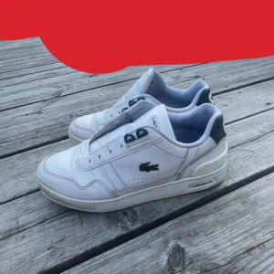 Vita Lacoste sneakers T-Clip - Snygga vita Lacoste T-Clip sneakers med gröna detaljer och klassisk krokodillogga på sidan. Skorna har snörning, platt sula och är tillverkade i skinn och syntet. Perfekta för dig som gillar en clean och sportig look.