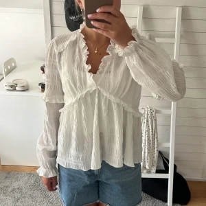 Vit omlottblus med volangdetaljer - Supersöt vit blus med omlottkänsla och volangdetaljer längs v-ringningen och ärmsluten. Lätt och luftig med lång ärm och struktur i tyget som ger en romantisk vibe. Perfekt att styla med jeans eller kjol för en trendig look.