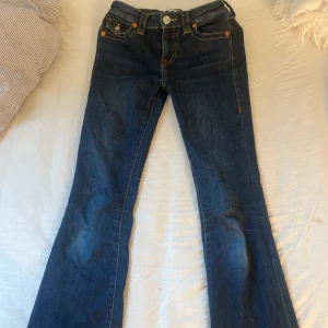 True Religion Becca Bootcut Jeans - Säljer ett par mörkblå True Religion Becca jeans med bootcut. Snygga kontrastsömmar, klassiska fickor med lock och logoknappar bak. Midwaist passform och tillverkade i jeansmaterial. Perfekta för dig som gillar en tidlös och cool look.