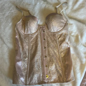Champagnefärgad korsett från Victoria's Secret - Superfin champagnefärgad korsett från Victoria's Secret med spetsdetaljer och satinpaneler. Den har bygelkupor, justerbara axelband och snygga hyskor framtill. Perfekt för dig som vill ha en lyxig och feminin look.
