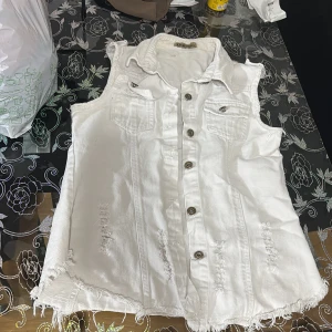 Vit jeansväst med slitna detaljer - Säljer en vit jeansväst med coola slitningar och fransad nederkant. Västen har två bröstfickor med knapp, klassisk krage och knäppning framtill. Perfekt för att ge outfiten en edgy touch.