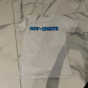 Vit Off-White t-shirt med blå logga - Lite oversized Vit t-shirt från Off-White med blå 3D-logga framtill och stora blå pilar på ryggen. Klassisk passform, rund hals och korta ärmar. Perfekt för dig som gillar streetwear och vill sticka ut med en ikonisk design.