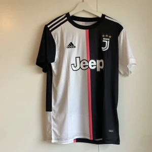 Juventus Adidas fotbollströja S - Juventus matchtröja från Adidas med svart och vit design, rosa rand i mitten och Jeep-logga på bröstet. Tröjan har korta ärmar, rund hals och tre vita ränder på axlarna. Tillverkad i lätt och ventilerande AEROREADY-material.