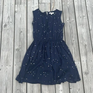 Mörkblå stjärnmönstrad klänning Hampton Republic - Supersöt mörkblå kortklänning från Hampton Republic med små guldiga  stjärnor över hela tyget. Klänningen är ärmlös med volangkant vid axlarna och har resår i midjan 
