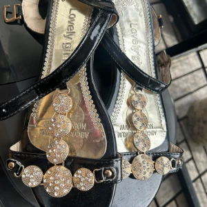 Svarta sandaler med guldiga detaljer - Glammiga svarta sandaler från Lovely girl med lackade remmar och snygga guldiga dekorationer med glittrande stenar framtill. Perfekta för dig som vill sticka ut med lite extra bling. Justerbart spänne på sidan och platt sula för skön känsla.