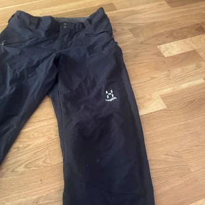 Svarta täckbyxor från Haglöfs herr S - Svarta täckbyxor från Haglöfs i herrmodell, storlek S. Byxorna har dragkedjor vid fickorna och är tillverkade i ett slitstarkt syntetmaterial, perfekta för utomhusaktiviteter under kyliga dagar. Klassisk design med broderad Haglöfs-logga på benet.