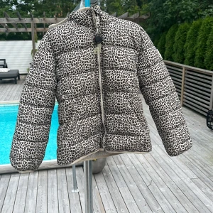Leopardmönstrad pufferjacka från GAP - Snygg pufferjacka från GAP med leopardmönster i beige och svart. Jackan har huva och är fodrad med mjuk, vit teddyfleece på insidan. Perfekt för dig som vill hålla dig varm under kalla dagar.