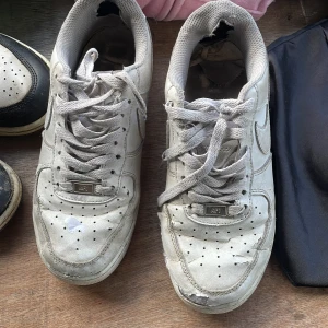 Nike Air Force 1 vita sneakers - Klassiska Nike Air Force 1 sneakers i vitt skinn med rund tå och snörning. Snygg streetstil med ikonisk Nike-logga på sidan och AIR-detalj på hälen. Perfekta för dig som gillar en clean och tidlös look