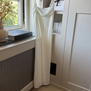 Vit långklänning från Gina Tricot - Elegant vit långklänning med tunna axelband och draperad urringning från Gina Tricot. Klänningen har en slät, mjuk yta och faller snyggt längs kroppen. Perfekt till sommaren!