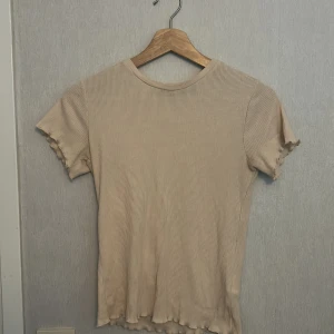 Beige ribbad topp från 157, stl S - En beige ribbad topp från 157 i storlek S. T-shirten har korta ärmar och en figurnära passform med vågiga kanter vid ärmslut och nederkant. Perfekt att styla med jeans eller kjol för en enkel och snygg look. Aldrig använt