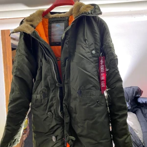 Grön parkajacka från Alpha Industries - Säljer en grön parkajacka från Alpha Industries med orange foder och fuskpäls vid huvan. Jackan har dragkedja, knappar, snörning i midjan och den klassiska 'Remove Before Flight'-taggen på ärmen. Perfekt för kalla dagar och riktigt snygg streetstil.