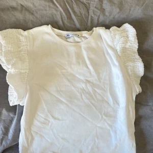 Vit volangtopp från Zara - Supersöt vit topp från Zara med ribbad struktur och stora volangärmar i smock. Perfekt för dig som vill ha något enkelt men ändå med en twist. Rund halsringning och ärmlös modell med extra volym på axlarna.
