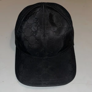 Svart keps från Gucci med mesh - Snygg svart keps från Gucci med klassiskt GG-monogram över hela framsidan. Kepsen har mesh-bakstycke och justerbar rem för perfekt passform. Materialet framtill är glansigt och ger en lyxig känsla, medan baksidan är i luftig mesh. Perfekt för dig som vill sticka ut. Köpte den hos en kille som köpte från sin vän då hans vän hade klippt av lappen i kepsen. Tveka inte hör av er vid intresse samt frågor.
