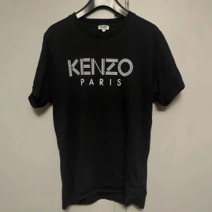 Svart Kenzo t-shirt med logga - Svart t-shirt från Kenzo med vit, grafisk logga på bröstet. Klassisk rund hals och korta ärmar. Tillverkad i mjuk bomull för en skön känsla. Perfekt för dig som gillar stilrena och trendiga plagg med statement-tryck.