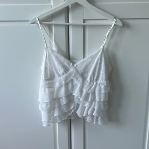 Vit volangig spetslinnetopp - Supersöt vit linnetopp med tunna axelband och flera lager volanger i spets och tyg. Toppen har v-ringning och en liten knappdetalj framtill 🤍 Använd 1 gång. Fler bilder kan fås