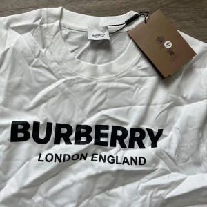 Vit Burberry t-shirt med logga - Vit t-shirt från Burberry med klassisk svart logga 'BURBERRY LONDON ENGLAND' tryckt framtill. T-shirten har rund halsringning och korta ärmar. Tillverkad i mjuk bomull för en clean och stilren look.